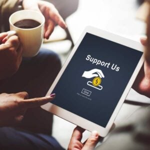 Two people sit together, one holding a tablet displaying a “Support Us” donation screen with a hand dropping a coin and a “Give” button. The other person holds a cup of coffee.